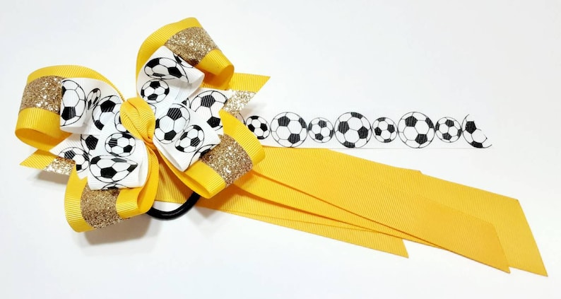 May include: A yellow and white hair bow with a soccer ball pattern. The bow has gold glitter accents and is attached to a black elastic hair tie. The bow is also accompanied by a yellow ribbon with a soccer ball pattern.