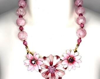 May include: A necklace featuring large, round, crackled light pink beads interspersed with smaller dark pink beads. The centerpiece consists of three enameled metal flowers in shades of white and pink, with the central flower featuring pink rhinestones.