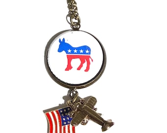 May include: A necklace featuring a circular pendant with a blue and red donkey graphic containing white stars. Attached to the pendant are two charms: a small metal airplane and a miniature American flag with red and white stripes and blue stars.