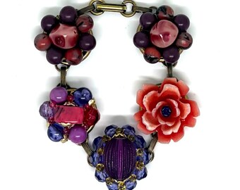 May include: A link bracelet featuring five distinct floral and beaded clusters in shades of purple, pink, and coral. The components are connected by antique brass-toned metal links, showcasing a variety of round, faceted, and carved flower-shaped beads.