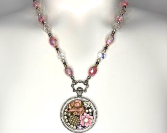 May include: A necklace featuring a silver-toned circular pendant filled with pink floral charms, small pearls, and clear rhinestones. The chain consists of alternating clear and iridescent pink faceted beads with small metallic spacer accents.