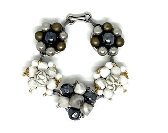 May include: A bracelet featuring five circular clusters of beads connected by silver-toned metal links. Two clusters contain dark grey and bronze beads, two contain white and marbled white beads, and one central cluster features dark grey and white beads.