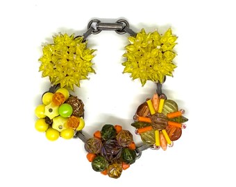 May include: A bracelet featuring a dark metal chain link base adorned with five distinct clusters of beads. The clusters include bright yellow starburst shapes, round yellow and green beads, and multi-colored orange, brown, and green faceted beads.