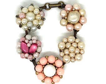 May include: A bracelet featuring five circular clusters of beads connected by bronze-toned metal links. The clusters include various shades of white, cream, and pink pearls, along with two smooth, oval-shaped hot pink cabochon beads.