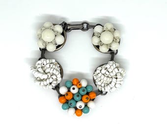 May include: A link bracelet featuring five circular clusters of beads. Two clusters contain white spherical beads, two contain white tubular beads, and one central cluster features a mix of white, orange, and teal spherical beads.