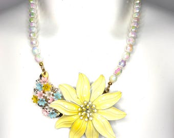 May include: A necklace featuring a large, pale yellow enamel flower pendant and a cluster of smaller pink, blue, and yellow enamel flowers. The chain consists of iridescent, faceted white beads with a subtle pearlescent finish throughout.