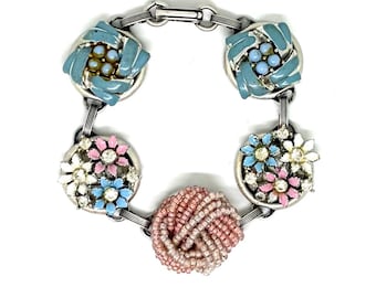 May include: A silver-tone link bracelet featuring five circular charms. Two charms have blue enamel and light blue beads, two have pink, blue, and white flower-shaped accents with clear rhinestones, and one is a spherical pink beaded knot.