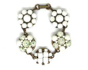 May include: A link bracelet featuring five circular brass settings connected by metal bars. Two settings contain white spherical beads in a floral arrangement, two contain a mix of white and green speckled beads, and one contains white square beads.
