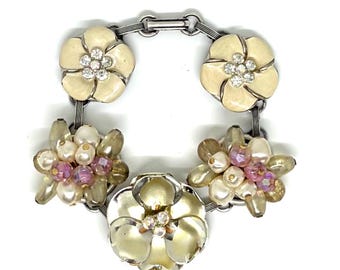 May include: A silver-toned link bracelet featuring two cream-colored enamel flowers with clear rhinestone centers, two clusters of cream and pink beads, and one large central cream-colored flower with a clear rhinestone center.