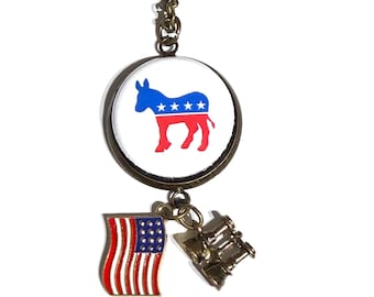May include: A bronze-toned necklace featuring a circular pendant with a blue and red donkey silhouette graphic containing white stars. Two smaller charms hang below: a waving American flag and a pair of bronze-colored binoculars.