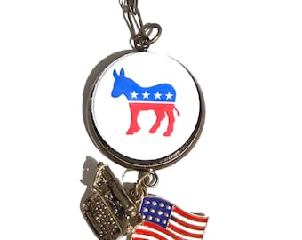 May include: A circular pendant on a bronze-toned chain featuring a white background with a blue and red donkey silhouette containing four white stars. Two smaller charms hang below: a metallic typewriter and a waving American flag enamel charm.
