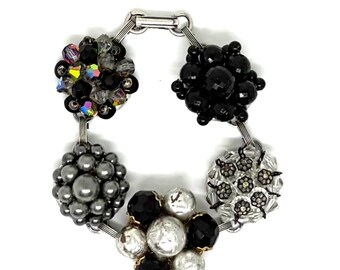 May include: A bracelet featuring five distinct clusters of beads connected by silver-toned metal links. The clusters include black faceted beads, iridescent crystals, metallic grey spheres, clear beads with small floral accents, and a mix of white and black beads.