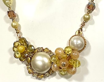 May include: A necklace featuring a central arrangement of a large cream-colored faux pearl surrounded by gold-toned leaves, a yellow faceted flower-shaped bead, and a cluster of translucent yellow and peach-colored beads on a gold-toned chain.