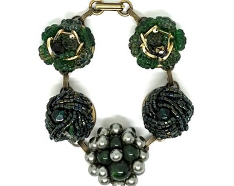 May include: A bracelet featuring five circular charms connected by gold-tone metal links. Two charms are made of green textured beads, two are composed of dark iridescent seed beads, and the central charm features silver and dark green beads.
