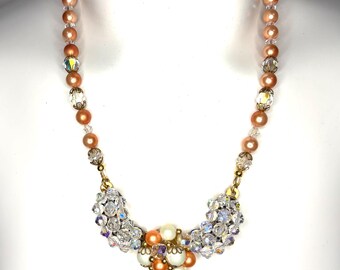 May include: A necklace featuring a strand of peach-colored round beads interspersed with clear faceted crystal beads. The center focal point consists of two clusters of clear crystals flanking a central arrangement of peach and cream-colored beads.