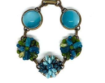 May include: A link bracelet featuring two smooth, round, translucent turquoise cabochons and three circular clusters of beads in shades of blue, teal, and olive green. The components are connected by antique brass-toned metal links and a clasp.