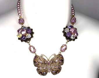 May include: A necklace featuring a central bronze-toned butterfly pendant adorned with small purple and clear crystals. The chain consists of light purple pearl beads, clear faceted beads, and clusters of purple beads set in bronze-toned metal.
