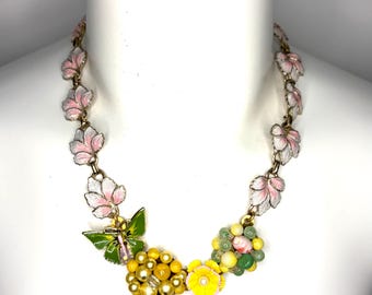 May include: A necklace featuring a chain of pink and white enameled leaf links. The centerpiece includes a green butterfly charm, a cluster of yellow beads, a single yellow flower with a white center, and a cluster of green and yellow beads.