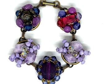 May include: A bracelet featuring five circular bronze-toned metal links connected by jump rings. Each link is adorned with clusters of beads in shades of purple, lavender, blue, and red, including faceted, smooth, and textured glass components.