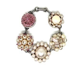 May include: A link bracelet featuring five circular charms connected by silver-toned metal bars. The charms include a faceted pink crystal, a central pearl surrounded by clear stones, a cluster of cream pearls, a large central pearl with smaller pearls, and a mix of translucent beads.