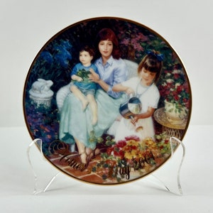 May include: A porcelain plate with a gold rim features a painting of a mother and her two children in a garden setting. The mother is wearing a light blue shirt and a white skirt. The children are wearing blue and white clothing. The plate has the text "Mother's Day 2001" on the bottom.