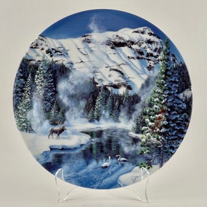May include: A white ceramic plate with a winter scene. The plate features a snowy mountain range, a frozen lake, a lone elk, and two swans. The plate is decorated with a blue and white glaze.