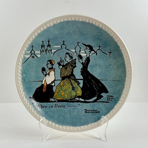 May include: A blue and white ceramic plate with a scene of four people walking in a procession. The plate has a white border with a beaded design. The scene is set against a blue background with white buildings and crosses. The text "When in Rome" is printed on the plate. The plate is on a white stand.