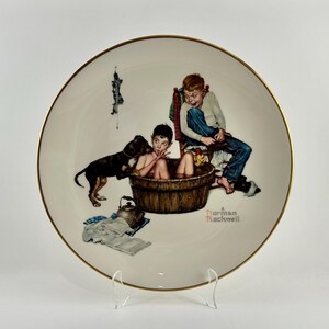 May include: A white porcelain plate with a gold rim features a Norman Rockwell painting of two boys and a dog. One boy is in a bathtub, the other is sitting on a chair, and the dog is standing next to the bathtub. The painting is titled "A Norman Rockwell".
