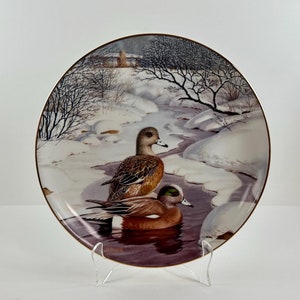 May include: A porcelain plate with a winter scene featuring two American wigeon ducks swimming in a stream. The plate has a gold rim and the artist's signature "JERNER" is visible in the bottom right corner.