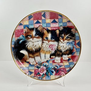 May include: A decorative ceramic plate featuring three kittens with colorful bows sitting on a patchwork quilt. The plate has a gold rim and is displayed on a white stand.