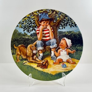 May include: A vintage ceramic plate featuring a painting of two children and a dog in a park setting. The boy is wearing a baseball cap and a red and white striped shirt. The girl is wearing a white dress and a white sun hat. The dog is brown and white and is sitting next to the boy. The plate has a green background with a tree and bushes.