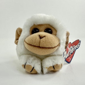 May include: White plush monkey with brown eyes and a smile. The monkey is wearing a white fur coat and has a red heart-shaped tag that says "Puffkins Collectible Key Rings".