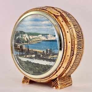 May include: A decorative barrel-shaped clock with a scenic image of a coastal town. The clock has a light brown, wood-like finish with detailed carvings and a silver-colored frame around the image. The clock stands upright on a small base.