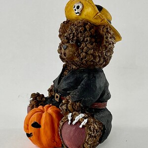 May include: A brown teddy bear wearing a black costume and a yellow pirate hat with a skull design. The bear is holding an orange jack-o'-lantern.