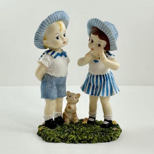 May include: A figurine of two children, a boy and a girl, wearing blue and white striped outfits and hats. They are standing on a green base with a small brown and white cat figurine between them.