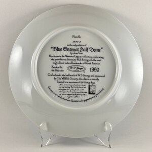 May include: A white ceramic plate with a blue and white image of a snow-capped mountain peak. The plate is part of a collection called "Nature's Legacy" and features the text "Blue Snow at Half Dome" by Jean Sian. The plate is numbered 10177-6 and is part of a limited edition of 150 firing days. The plate is crafted under the hallmark of W.S. George and sponsored by The Wildlife Society.