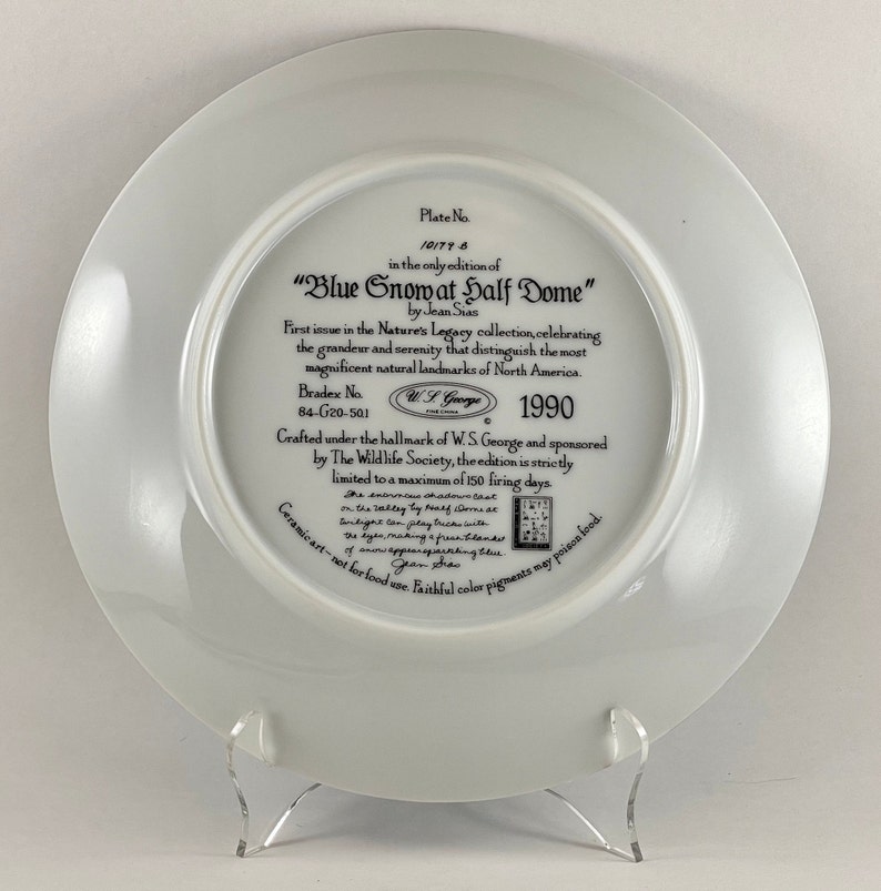 May include: A white ceramic plate with a blue and white image of a snow-capped mountain peak. The plate is part of a collection called "Nature's Legacy" and features the text "Blue Snow at Half Dome" by Jean Sian. The plate is numbered 10177-6 and is part of a limited edition of 150 firing days. The plate is crafted under the hallmark of W.S. George and sponsored by The Wildlife Society.