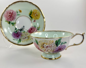 Peut inclure: Une tasse à thé et une soucoupe délicates avec un motif floral. La tasse et la soucoupe sont blanches avec un fond vert clair et présentent un motif floral de chrysanthèmes roses, jaunes et blancs. Les bords de la tasse et de la soucoupe sont bordés d'or.