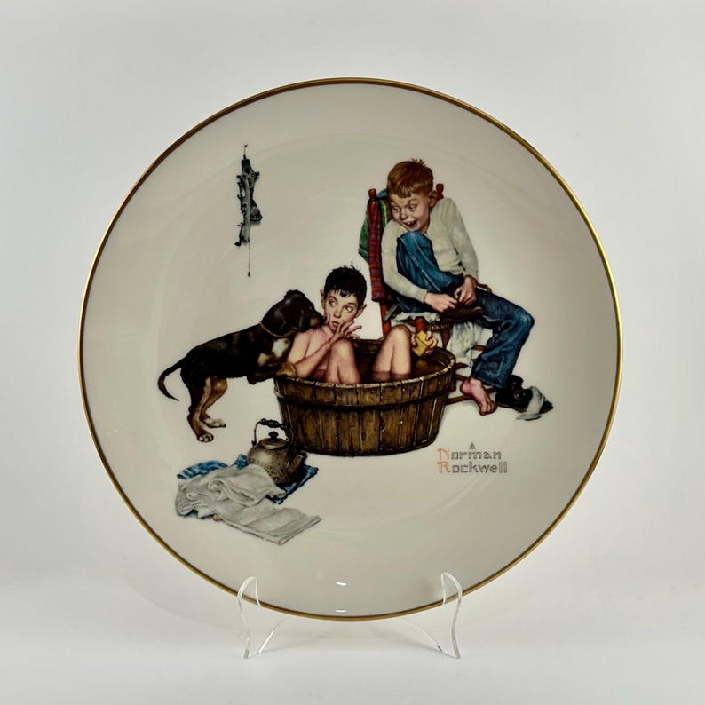 May include: A white porcelain plate with a gold rim features a Norman Rockwell painting of two boys and a dog. One boy is in a bathtub, the other is sitting on a chair, and the dog is standing next to the bathtub. The painting is titled "A Norman Rockwell".