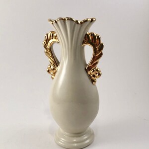 May include: A white ceramic vase with gold leaf accents and two handles. The vase has a fluted design and a rounded base.