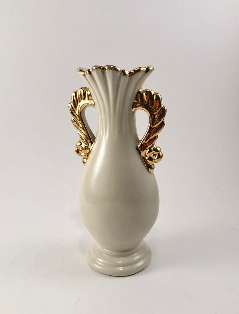 May include: A white ceramic vase with gold leaf accents and two handles. The vase has a fluted design and a rounded base.