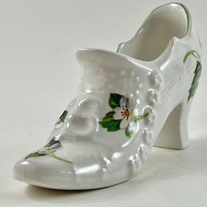 May include: White ceramic shoe with a floral design. The shoe has a high heel and is decorated with green leaves and white flowers.