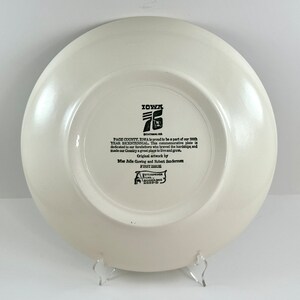 May include: A white ceramic plate with a black and white design commemorating the 200th anniversary of Page County, Iowa. The plate features the text "IOWA 76 BICENTENNIAL", "PAGE COUNTY, IOWA is proud to be a part of our 200th YEAR BICENTENNIAL. This commemorative plate is dedicated to our forefathers who braved the hardships and made our Country a great place to live and grow." and "Original artwork by Bliss Julie Gowing and Robert Sunderman FIRST ISSUE".
