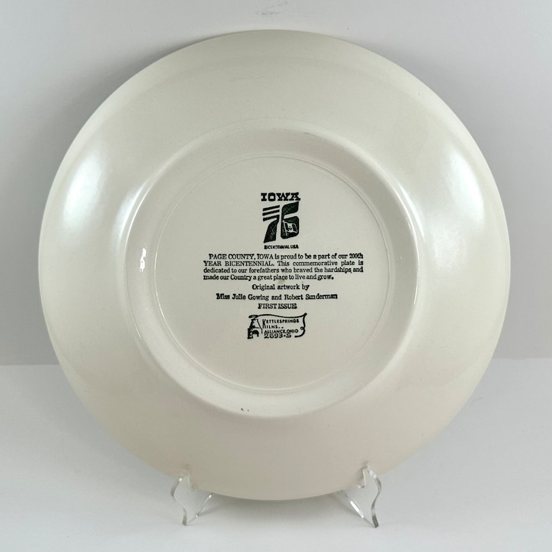 May include: A white ceramic plate with a black and white design commemorating the 200th anniversary of Page County, Iowa. The plate features the text "IOWA 76 BICENTENNIAL", "PAGE COUNTY, IOWA is proud to be a part of our 200th YEAR BICENTENNIAL. This commemorative plate is dedicated to our forefathers who braved the hardships and made our Country a great place to live and grow." and "Original artwork by Bliss Julie Gowing and Robert Sunderman FIRST ISSUE".