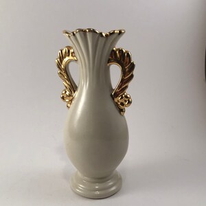 May include: A white ceramic vase with gold leaf handles. The vase has a fluted design and a round base.