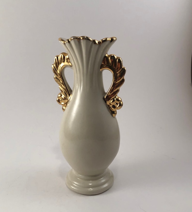 May include: A white ceramic vase with gold leaf handles. The vase has a fluted design and a round base.