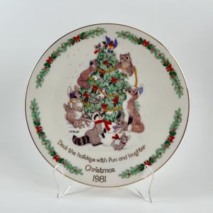 May include: A decorative Christmas plate featuring a festive scene with woodland animals gathered around a decorated Christmas tree. The plate has a gold rim and a border of green foliage and red bows. Text on the plate reads "Deck the holidays with fun and laughter Christmas 1981."
