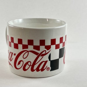 Op de afbeelding: Witte keramische mok met het Coca-Cola-logo in rood schrift. De mok heeft een rood-zwart geblokt patroon rond de bovenkant. De mok heeft een handvat en is ontworpen voor dranken.
