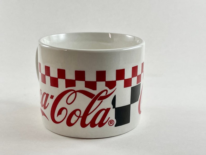 Op de afbeelding: Witte keramische mok met het Coca-Cola-logo in rood schrift. De mok heeft een rood-zwart geblokt patroon rond de bovenkant. De mok heeft een handvat en is ontworpen voor dranken.