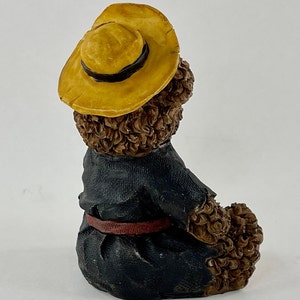 May include: A brown teddy bear figurine wearing a yellow straw hat with a black band and a blue jacket with a red belt. The bear is sitting with its back to the camera.